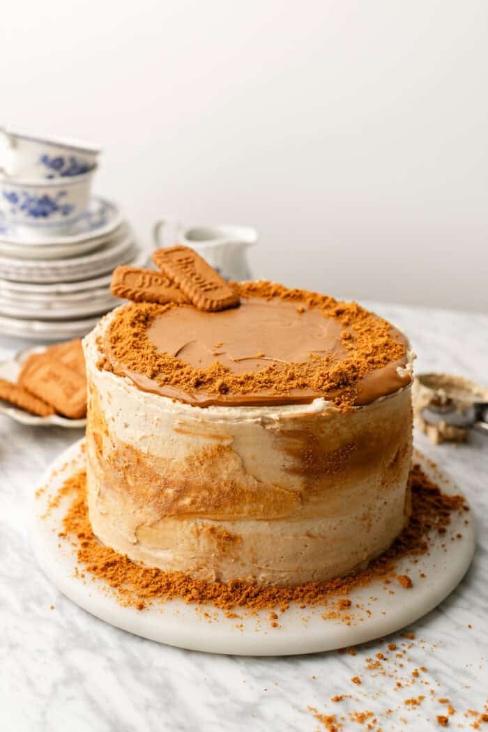 A round cake with light brown frosting is topped with cookie spread and two rectangular cookies, with cookie crumbs scattered on top and around the base. Plates and teacups are in the background.