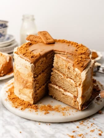 A three-layer cake with tan frosting, biscuit crumbs on top and around, and two whole biscuits on top, sits on a marble surface with a slice removed. Plates and a milk bottle are in the background.
