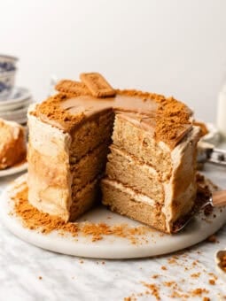 A three-layer cake with light brown frosting and crumbs, topped with rectangular cookies. A large slice has been cut out and sits on a marble surface with scattered crumbs.