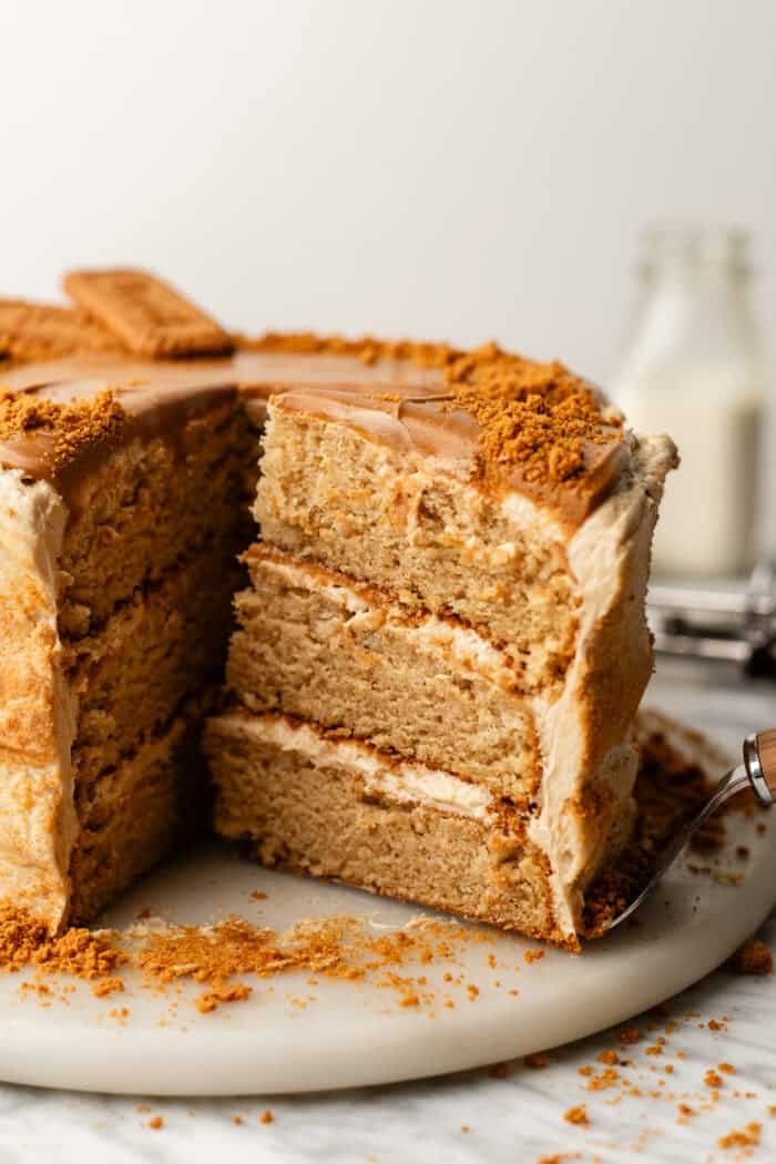 A three-layer cake with brown frosting and biscuit crumbs on top, missing one slice. A cake server is under the removed slice. A bottle of milk is in the background.