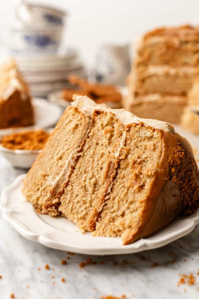 A close-up of a three-layer slice of cake with light brown frosting, served on a white plate, with crumbs scattered around and more cake and dishes in the blurred background.