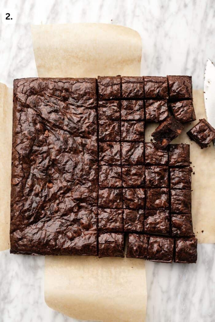 full batch of brownies with half of it sliced into bite sized pieces and the other half remaining whole on a parchment paper piece on a marble table.