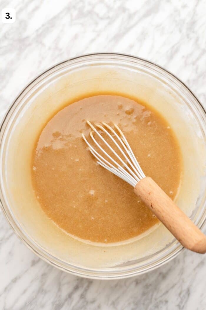 Bowl with a whisk whisking the chocolate cake batter before the cocoa powder is added in on a marble table.