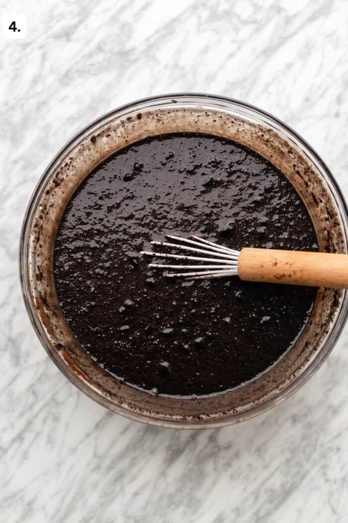 Bowl with a whisk whisking the chocolate cake batter with the cocoa powder added in on a marble table.
