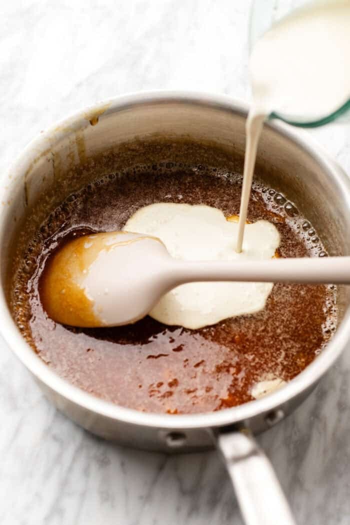 pouring in the vegan heavy cream into a saucepan over medium heat on a marble table.