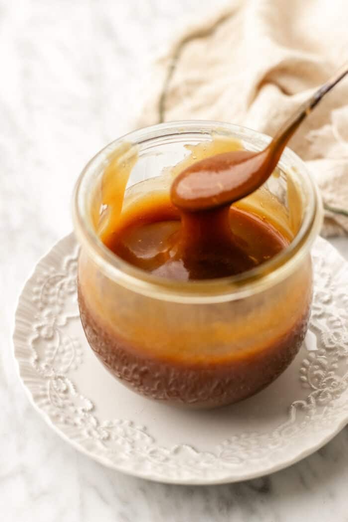 thickened caramel sauce in a jar on a white plate on a marble table with a spoon dripping the caramel from the jar.