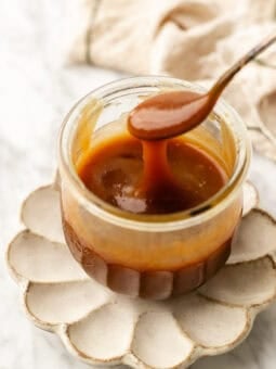 jar of vegan caramel sauce with a spoon drizzling the caramel on a flower petal plate with a cloth towel in the background on a marble table.