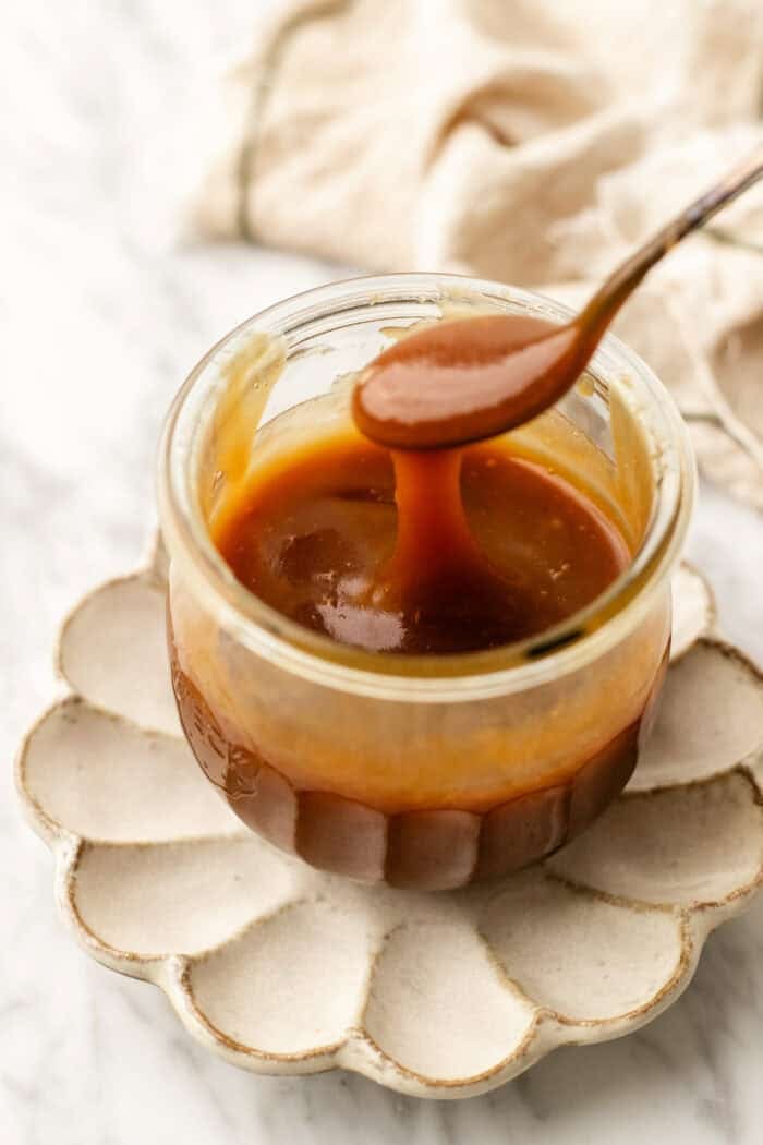 jar of vegan caramel sauce with a spoon drizzling the caramel on a flower petal plate with a cloth towel in the background on a marble table.