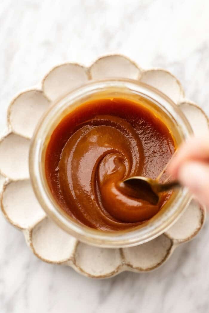 thickened caramel in a jar on a petal plate with the spoon swirling it around on a marble table.