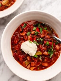 A bowl of chili topped with a dollop of sour cream and chopped herbs, with a spoon in the bowl, on a marble surface. Another bowl of chili is partially visible in the background.