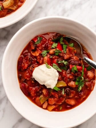 A bowl of chili topped with a dollop of sour cream and chopped herbs, with a spoon in the bowl, on a marble surface. Another bowl of chili is partially visible in the background.