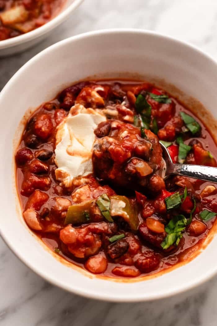 A bowl of chili with beans, vegetables, and tomatoes, topped with a dollop of sour cream and chopped herbs, with a spoon in the bowl.