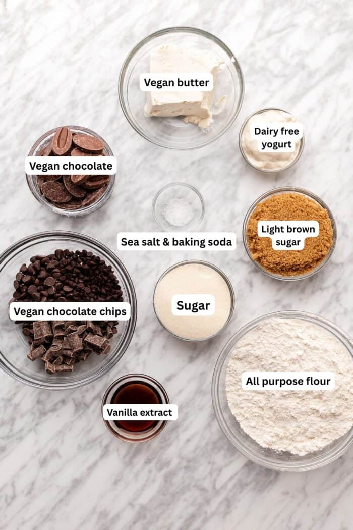 Bowls with labeled baking ingredients on a marble surface: vegan butter, dairy-free yogurt, vegan chocolate, sea salt & baking soda, light brown sugar, sugar, vegan chocolate chips, vanilla extract, and all-purpose flour.