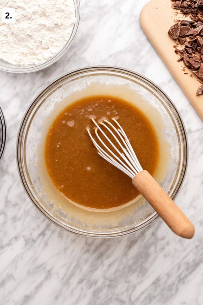 A glass bowl with a whisk mixing a brown liquid batter sits on a marble surface, surrounded by a bowl of flour and a cutting board with chopped chocolate.
