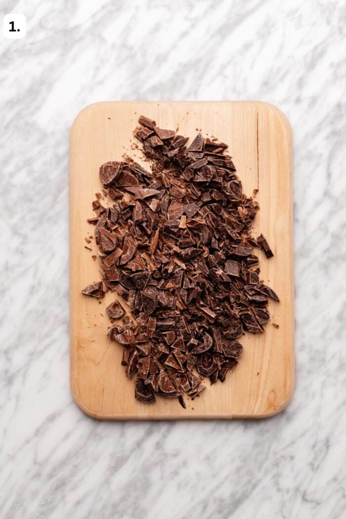 Chopped chocolate pieces on a wooden cutting board, placed on a white marble surface.