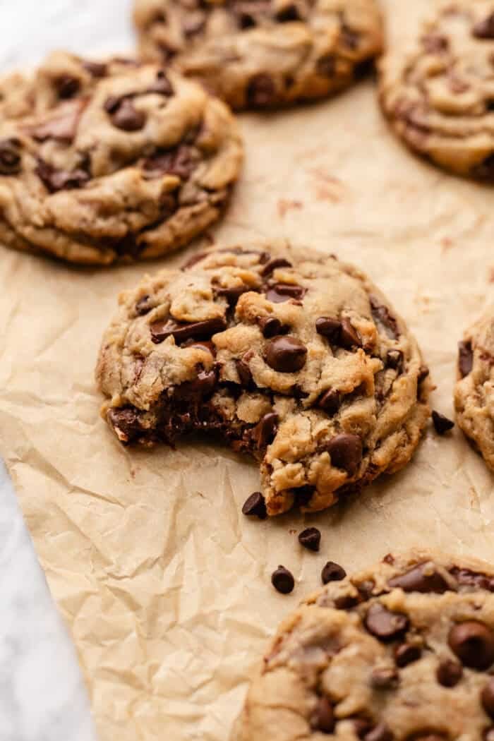 A chocolate chip cookie with a bite taken out of it sits on parchment paper, surrounded by other whole cookies.