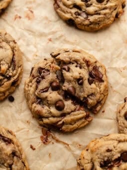 A close-up of chunky chocolate chip cookies on crinkled parchment paper, with melted chocolate chips visible in the gooey, golden-brown cookies.