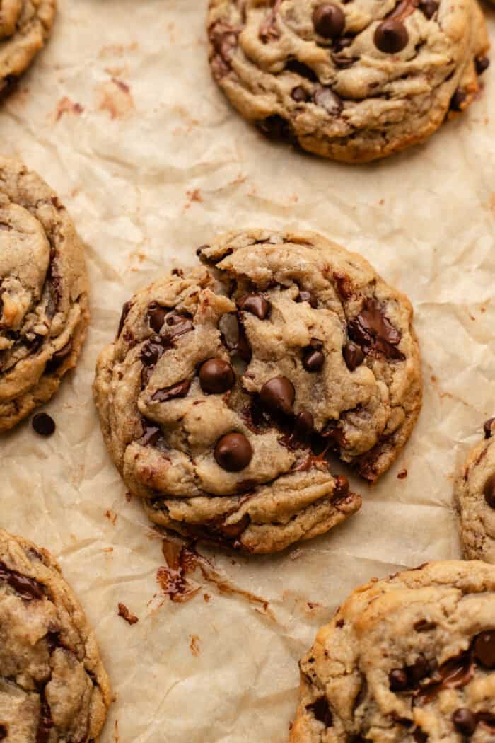 A close-up of chunky chocolate chip cookies on crinkled parchment paper, with melted chocolate chips visible in the gooey, golden-brown cookies.