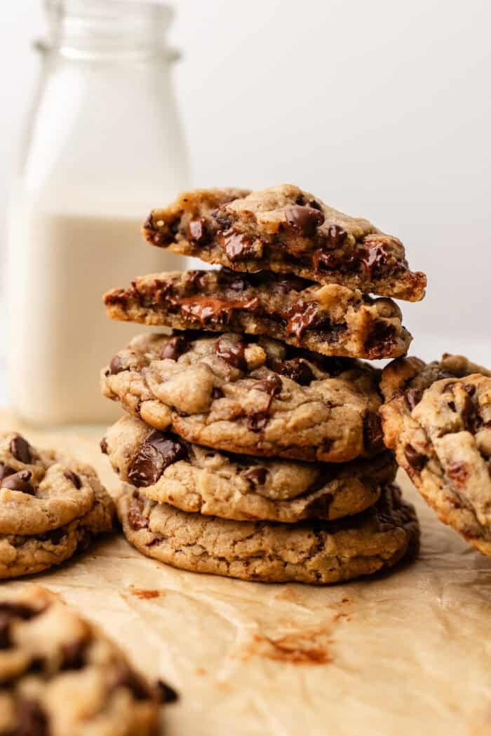 A stack of chocolate chip cookies sits on parchment paper, with a glass bottle of milk in the background. Some cookies are broken in half, showing the gooey chocolate chips inside.
