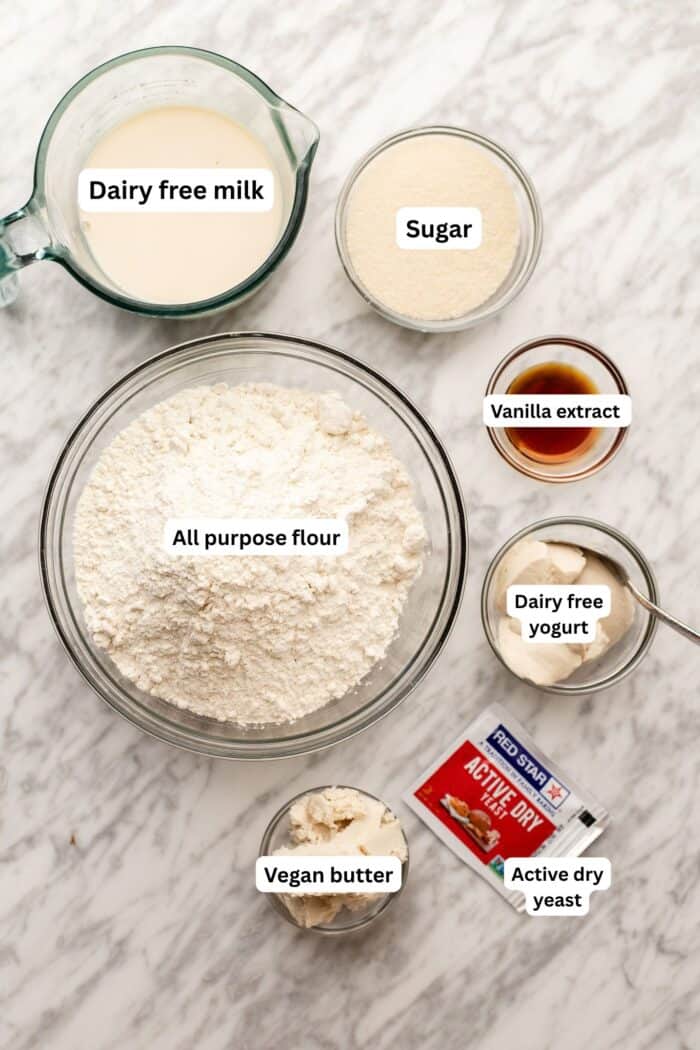 Bowls of dairy free milk, sugar, all purpose flour, vanilla extract, dairy free yogurt, vegan butter, and a packet of active dry yeast are arranged on a marble surface, each labeled with text indicating their contents.