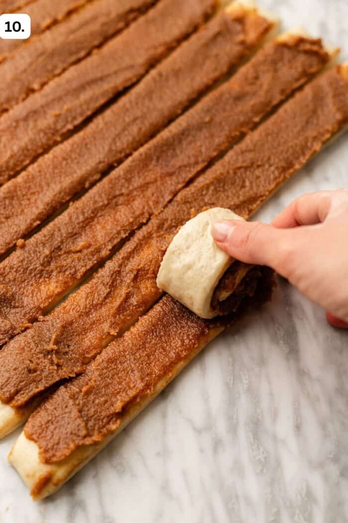A hand rolls a strip of dough covered with cinnamon filling on a marble surface. Several unrolled strips with cinnamon spread are laid out next to it.