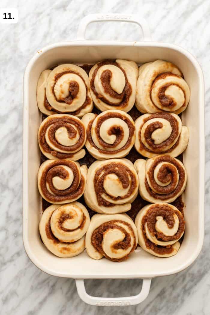 A white baking dish filled with 12 unbaked cinnamon rolls placed close together on a marble surface.