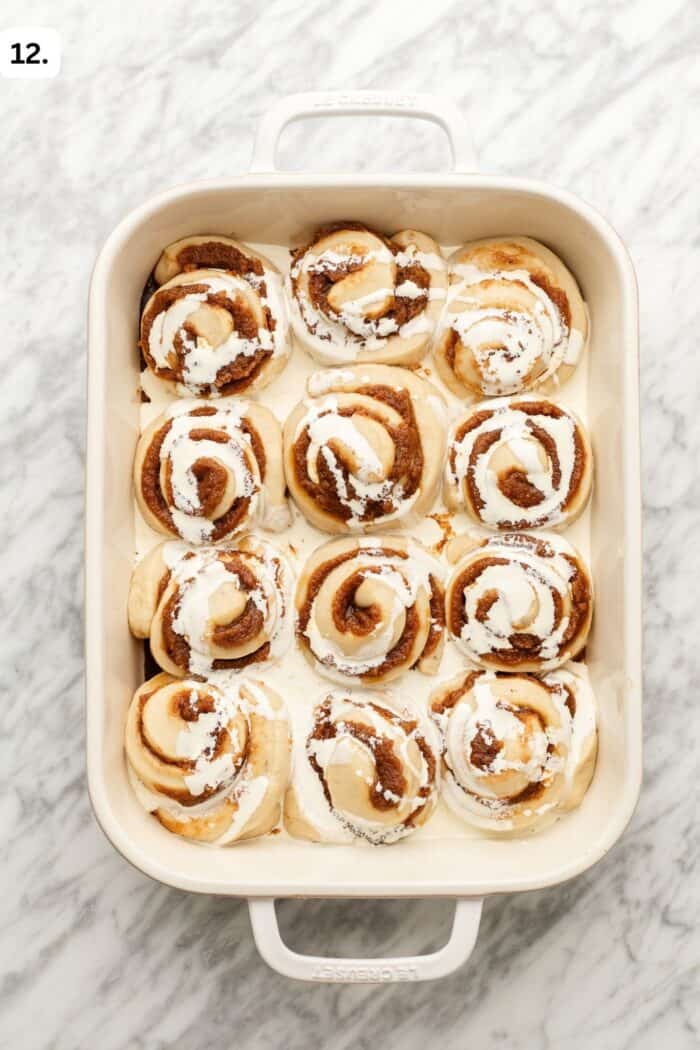 A baking dish with 12 cinnamon rolls topped with swirled white icing, set on a marble countertop.