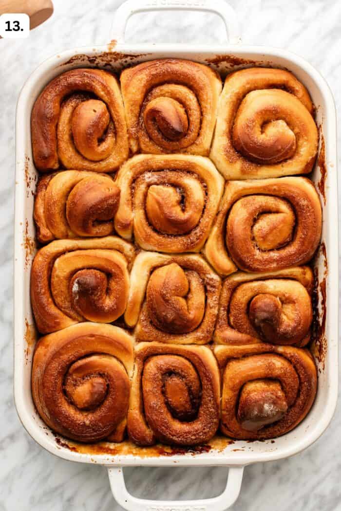 A white baking dish filled with twelve golden-brown cinnamon rolls arranged in rows, sitting on a marble surface.