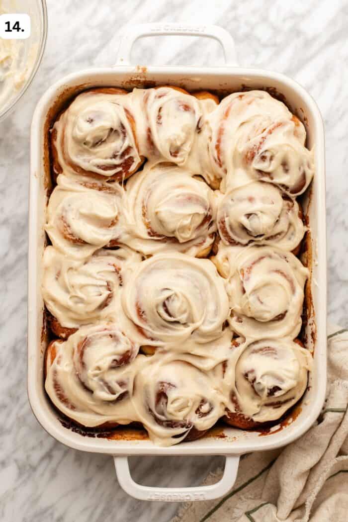 A white baking dish filled with twelve frosted cinnamon rolls sits on a marble countertop, partially wrapped in a towel, with creamy icing swirled on top of each roll.
