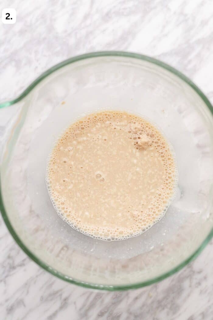 Glass bowl with bubbly, light brown liquid mixture on a marble surface, viewed from above. The liquid appears frothy and partially settled, suggesting an ingredient being activated or mixed. A number 2. is in the top left corner.