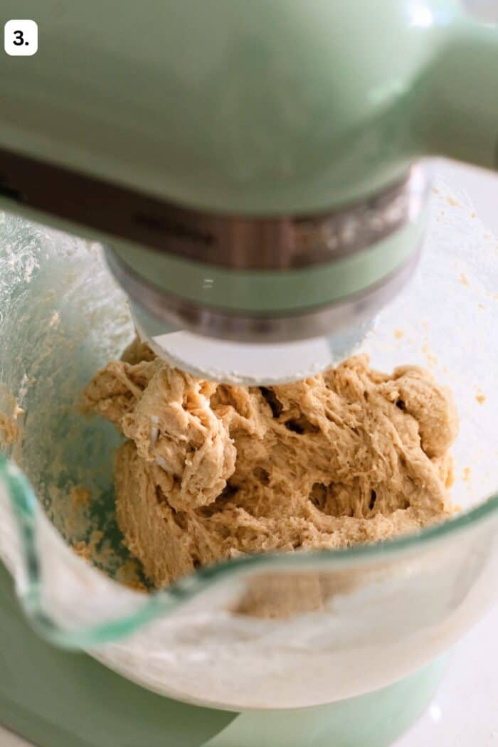 A stand mixer with a glass bowl is mixing a thick, beige dough. The bowl is partially filled, and the dough appears to be in the process of being kneaded. A small 3. is in the top left corner.