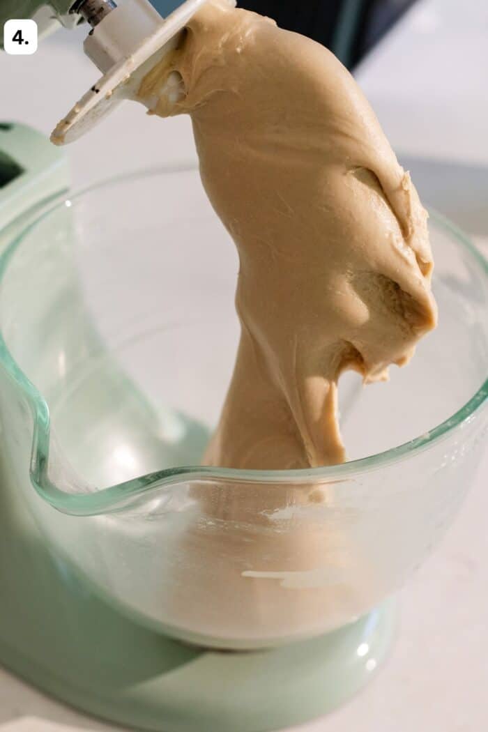 A close-up of bread dough being mixed in a stand mixer with a glass bowl. The dough is smooth and stretchy, clinging to the paddle attachment.
