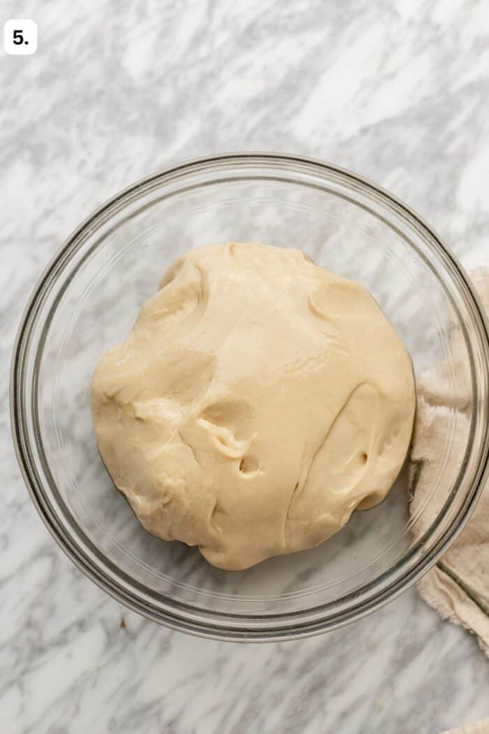 A glass bowl filled with smooth, risen dough sits on a marble surface. A beige cloth is partially visible at the edge.