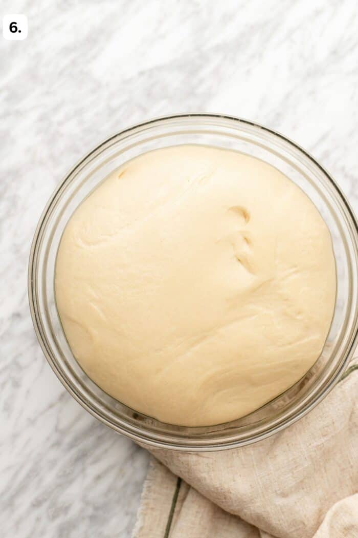 A glass bowl filled with risen dough sits on a light marble surface, partially on top of a beige cloth. A small number six is in the top left corner of the image.