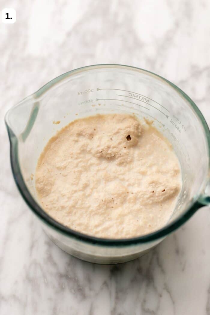 A glass measuring cup filled with bubbly yeast mixture sits on a marble surface. The liquid is creamy with some foam and bubbles on top. A number 1 is in the top left corner of the image.