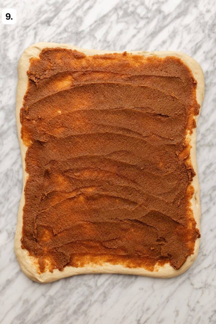 Rectangular rolled-out dough with a thick, even layer of brown cinnamon filling spread on top, sitting on a light marble surface. A small label with the number 9 is in the top left corner.
