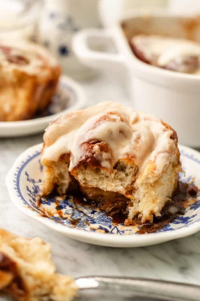 A cinnamon roll with icing is on a blue and white plate, partially eaten. More cinnamon rolls in a white dish and another plate are blurred in the background.