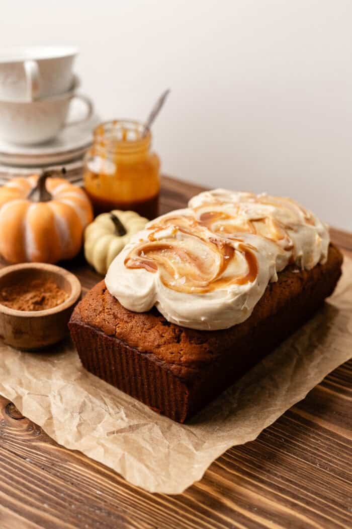 frosted pumpkin loaf with a drizzle of caramel on top and a jar of caramel behind the loaf with mini pumpkins next to it.