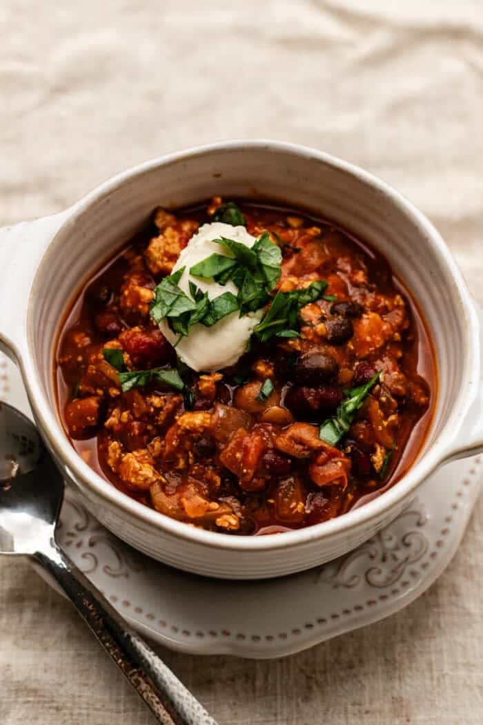 bowl of vegan pumpkin chili with sour cream on top on a plate with a cloth napkin underneath.