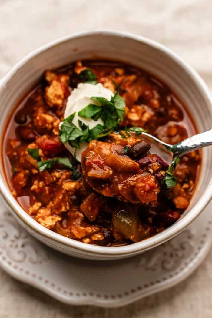 close up of a bowl of pumpkin chili, the spoon scooping the chili out with sour cream and herbs on top.