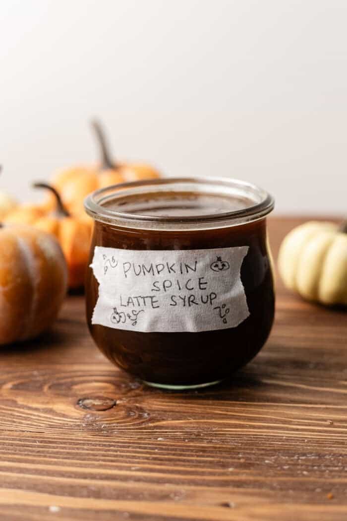 A glass jar of dark pumpkin spice latte syrup with a handwritten label, surrounded by small pumpkins on a wooden table.