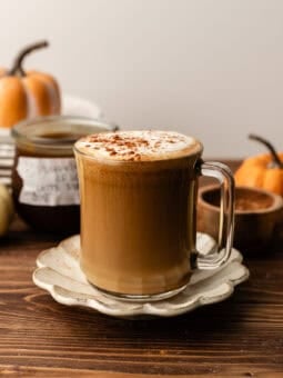 A glass mug of latte topped with foam and spice, resting on a scalloped saucer on a wooden table.