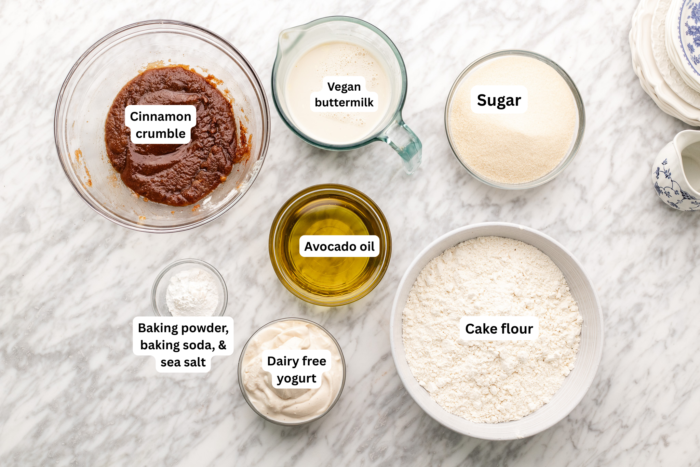 Ingredients for a recipe arranged on a marble surface: cinnamon crumble, vegan buttermilk, sugar, avocado oil, baking powder, baking soda, sea salt, dairy-free yogurt, and cake flour.