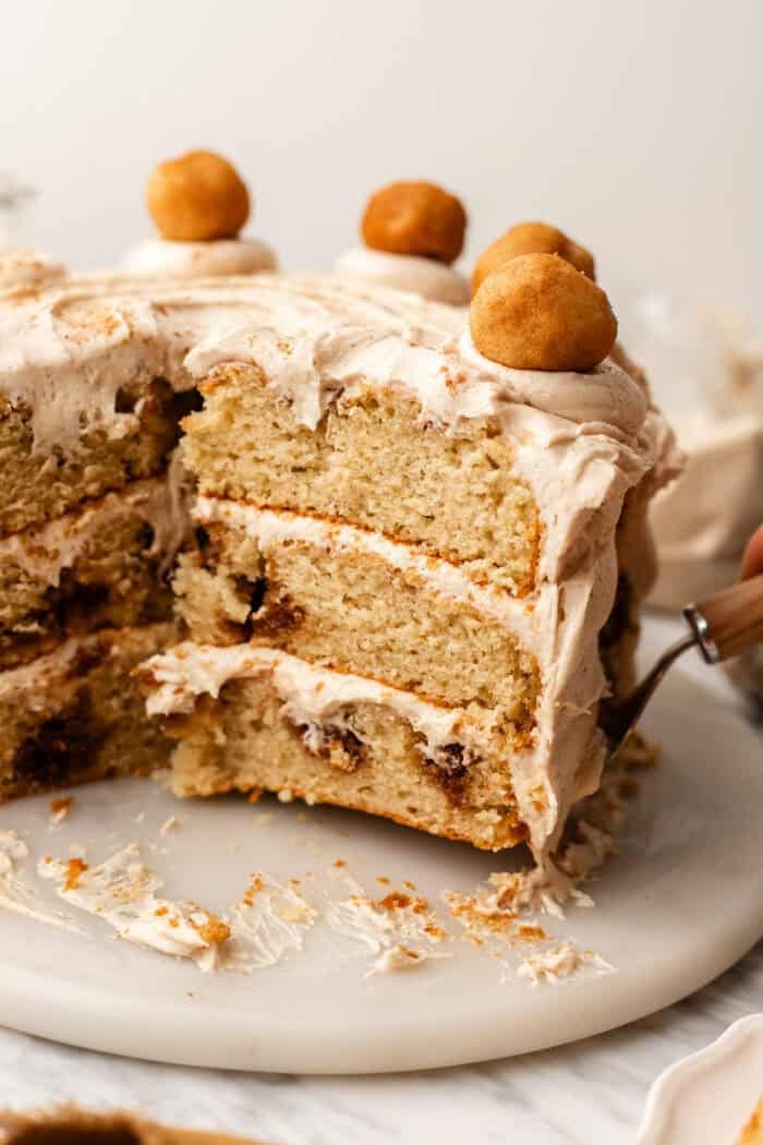 A three-layer cinnamon cake with cream frosting and small doughnut-like toppings, with a slice being removed.