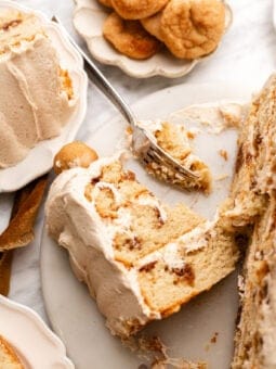 A slice of cinnamon roll cake with cream cheese frosting on a plate, with a fork and a bowl of snickerdoodle cookies in the background.