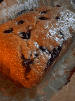 A golden-brown lemon blueberry loaf cake with visible blueberries, dusted lightly with powdered sugar, sits on a sheet of crinkled parchment paper.