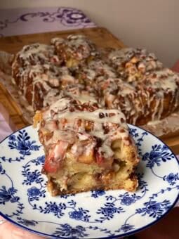 A square piece of apple fritter cake with icing on a blue and white patterned plate, with the rest of the pull-apart loaf in the background.